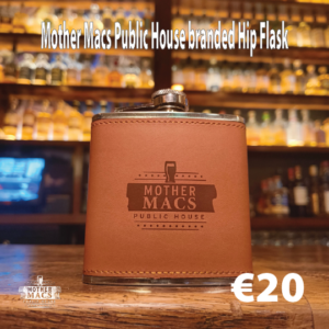 A brown hip flask branded with the logo Mother Macs Public house sitting on a bar counter infront of whiskey bottle back drop